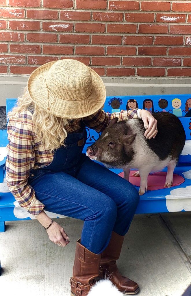 A woman dressed in a plaid shirt, tall brown boots, denim overalls and wearing a straw hat sits on a bench and pets a black and white pig.