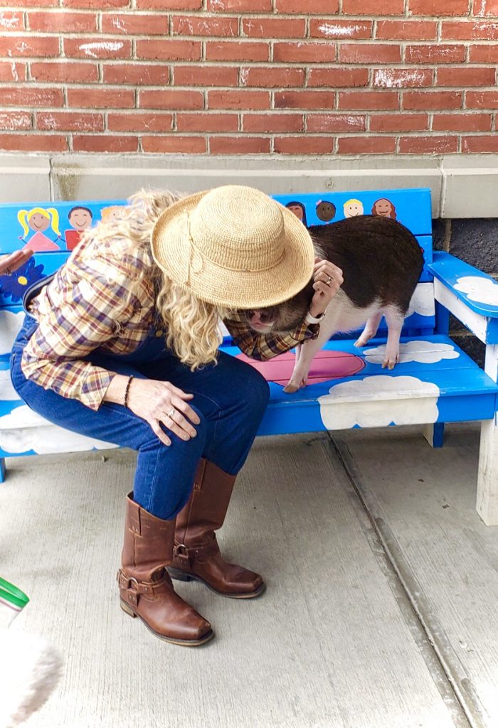 A woman dressed in a plaid shirt, tall brown boots, denim overalls and wearing a straw hat sigs on a bench and kisses a black and white pig.
