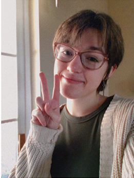 a high school girl with short hair and glasses holds up a v for victory (and peace). She is wearing a green shirt and beige sweater and she is smiling.