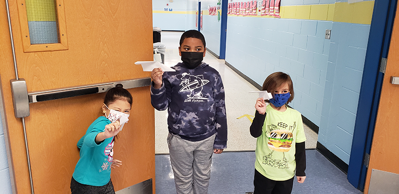 Three students, one girl on the left, and two boys,all wearing masks, hold up their paper airplanes. All are wearing masks.