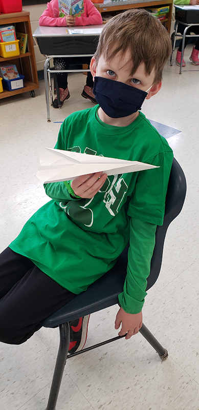 A boy wearing a green shirt and black mask sits in a chair and holds up his paper airplane.