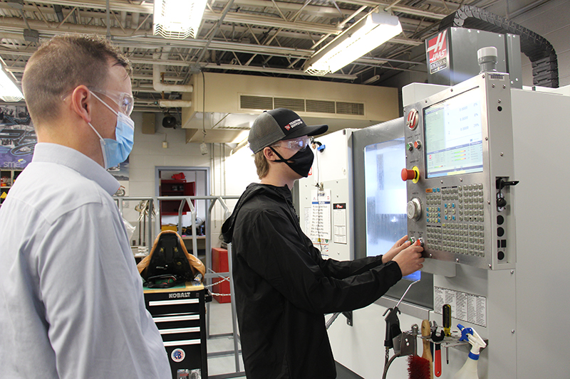 A young man in a black hoodie, hate and mask stands in front of a large gray machine with his hands on the controls. A man with short hair, button down shirt and blue mask stands behind him watching. They are in a workshop.