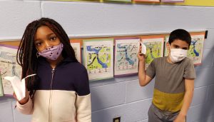 Two students, a girl and a boy, hold up their paper airplanes. Each is wearing a mask. The girl is on left and has long dark hair and is wearing a two-color sweatshirt. The boy is at right and wearing a gray and tan short-sleeve shirt.