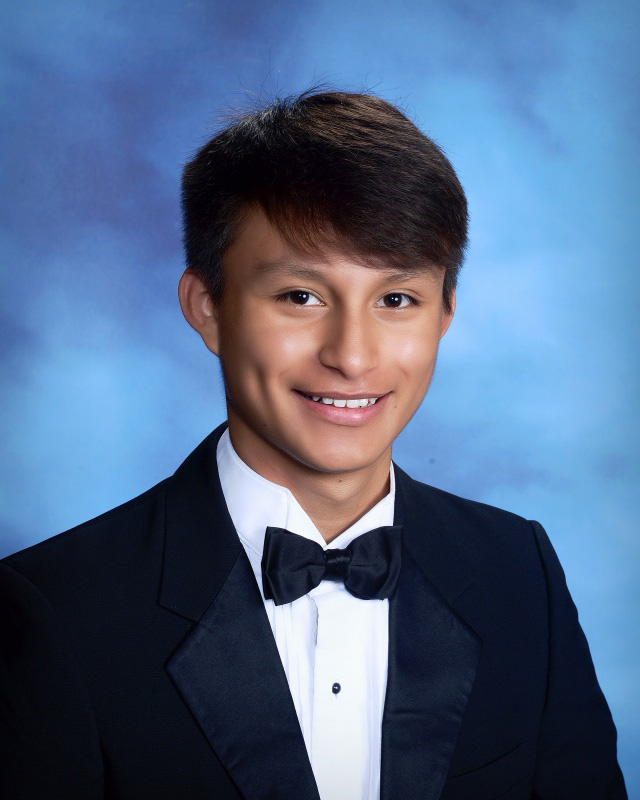 A young man in a black tuxedo, short dark hair, smiling. A blue background.