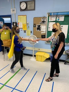 Two women in a classroom. The one on the left is wearing a blue shirt that says Teachers are just super heroes in disguise and a gold cape. She hands a gift to a woman on the right who has long blonde hair and  is wearing a black shirt. Both are wearing face masks.