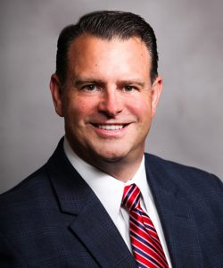 A man smiling with dark hair, wearing a white shirt and red striped tie and a blue suit jacket.