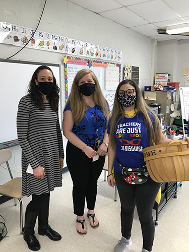 Three women stand in front of a classroom, whiteboards in back.  The woman on the right has long hair and is holding a wicker baskete. She is wearing a blue shirt that says Teachers are just super heroes in disguise. Next to her are two women, both with long hair. One is wearing a striped dress the other a blue shirt and black pants. They are all wearing face masks.