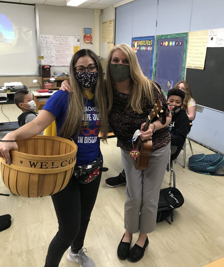 A woman with long blonde hair, wearing a dark shirt and gray pants has her arm around a woman with long hair, holding a wicker basket