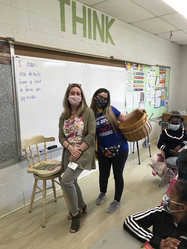 Two women stand in front of a classroom, with a white board in the background and the word THINK in large letters above it. Woman on the right holds a wicker basket and a blue shirt that says Teachers are just super heroes in disguise. The woman on the left has long blonde hair and is wearing gray pants and sweater. Both are wearing masks.