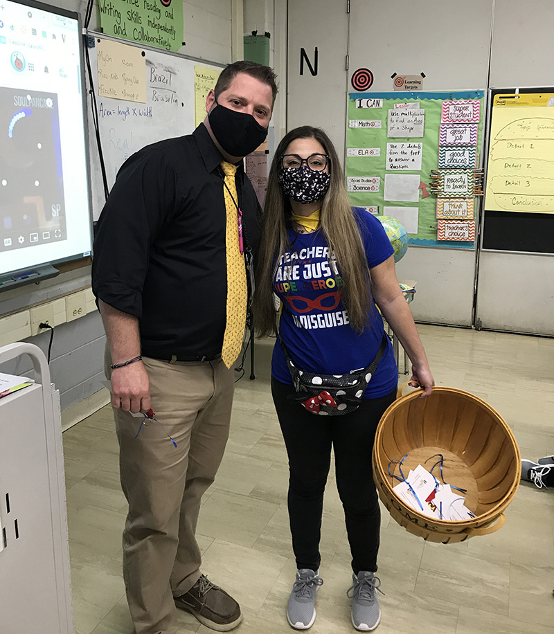 A man in a black shirt and gold time wearing a black facemask stands with a woman with a wicker basket, blue shirt that says Teachers are just super heroes in disguise. She is wearing a mask too. They are in a classroom.