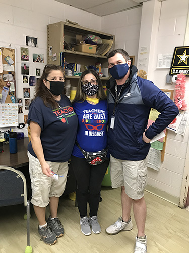 A woman in a blue shirt that says Teachers are just super heroes in disguise stands between a man on the right in a black shirt and tan shorts and a woman on the left also wearing a black shirt and tan shorts. All are wearing facemasks.