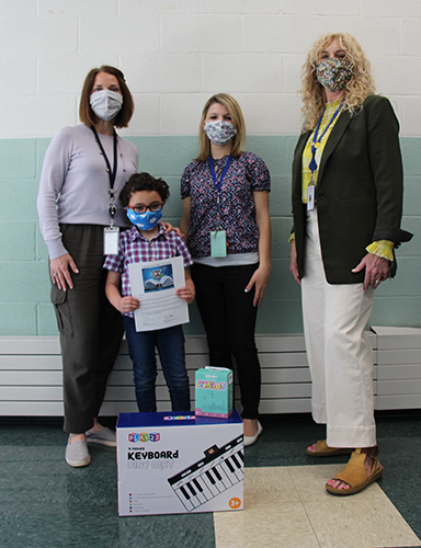 A little boy wearing a plaid shirt stands holding a certificate. Three women are around him, one with a gray shirt and chin-length arker hair, one with a flowered shirt and blonde hair, the third with longer blonde wavy hair wearing a dark jacket, yellow shirt and white pants. There is a box in front with  a picture of a keyboard on it. They all have masks on.