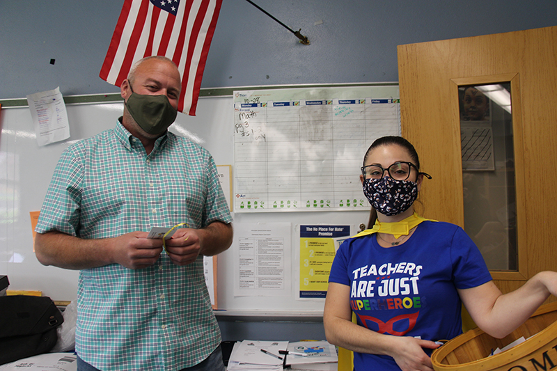 A woman in a blue t-shirt that says Teachers are just super heroes in disguise, wearing a gold cape and a face mask, stands with a man in a green shirt, wearing a facemask and holding a key chain she just gave him as a gift. There is an American flag behind them.