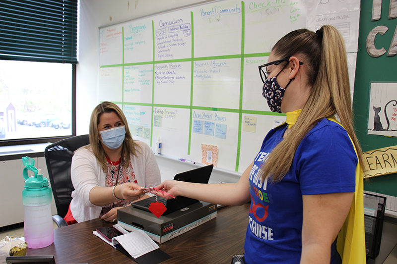 A woman sits at a desk with a computer in front of her. She is wearing a white sweater and a blue facemask. She has shoulder-length blonde hair. A woman in a blue shirt and long ponytail hands her a key chain as a gift. 