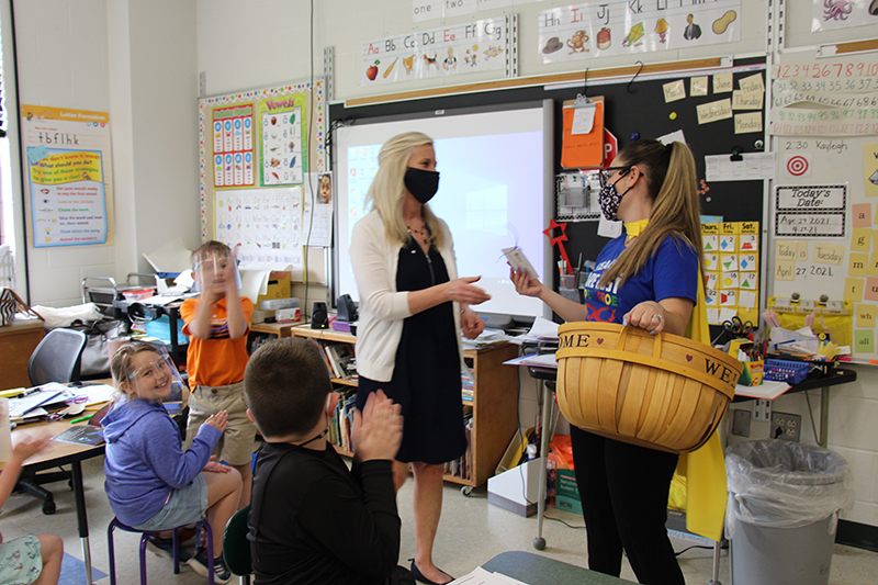 Two women stand in front of a classroom. Thewoman on the right is holding a wicker basket in one hand and a key chain in the other. She has a long ponytail. The woman on the left is wearing a black dress and white sweater. She has long blonde hair and is wearing a face mask. She has her hand out to accept the key chain gift. Students are watching and smiling and clapping.