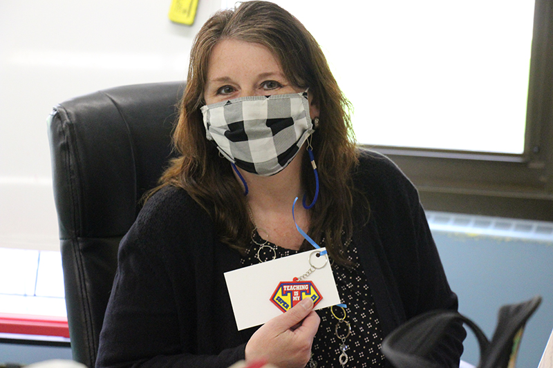 A woman wearing a black shirt and black plaid face mask holds up her key chain that says Teaching is my super power.
