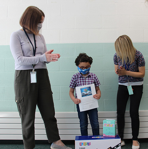 A little boy wearing a plaid shirt holds a piece of paper while two women, one on either side of him, clap. All are wearing masks.