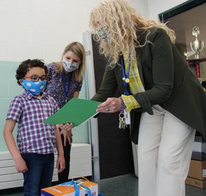 A woman with long blonde hair holds a green folder and shows it to a little boy wearing a plaid shirt. In the bak is a woman with shoulder-length blonde hair leaning in talking to him.