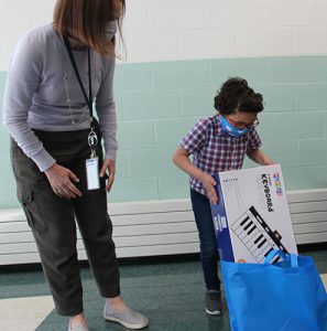 A little boy wearing a plaid shirt pulls a box out of a blue bag. The box has a keyboard on it. A woma wearing a gray sirt and dark pants is watching.