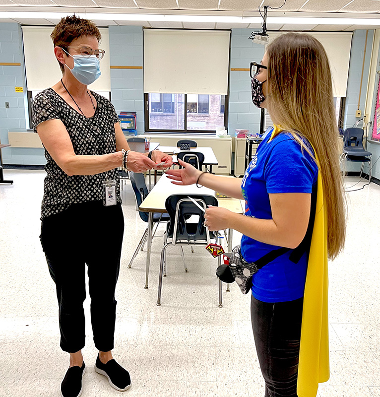 Two women in a classroom. Woman on the right has long hair and is wearing  a mask and glasses, blue t-shirt and gold cape. She is handing a gift - a key chain - to the woman on the left, who has short brown hair and is wearing a blue mask and shortsleve shirt, black pants.