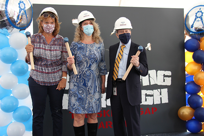 Three people - two women and one man - stand in front of a gray background. They are wearing hard hats and holding large hammers as if to begin construction. All are wearing masks