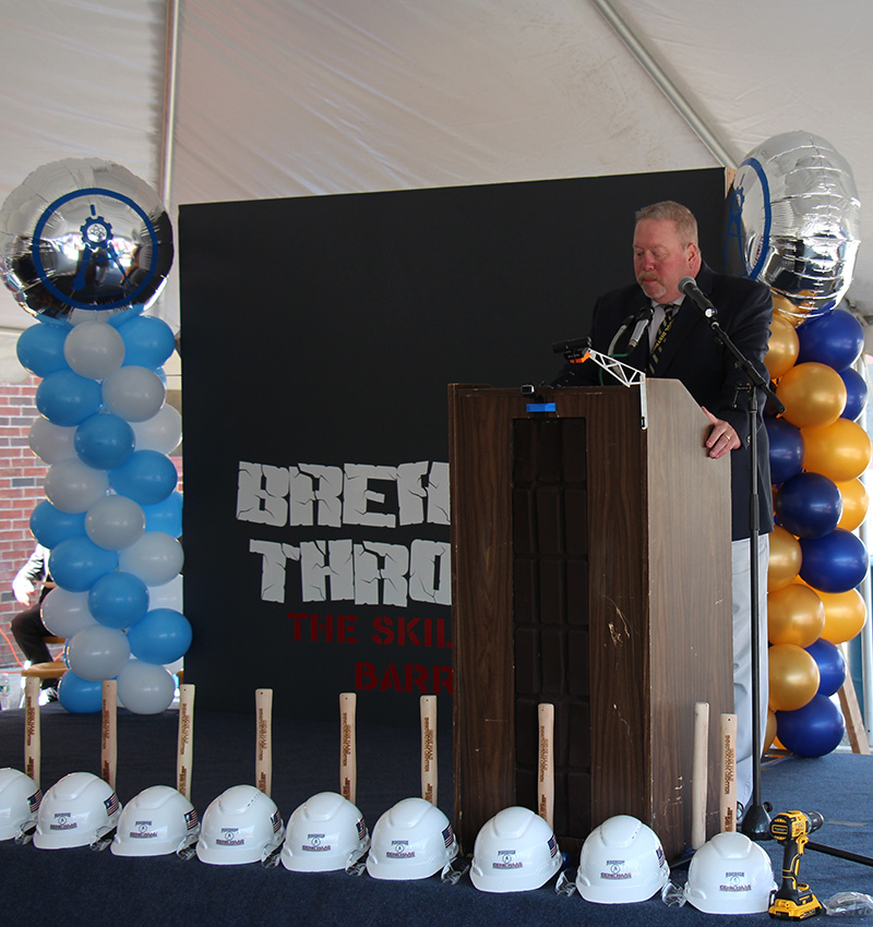 Hard hats and hammers line a stage where a man in a suite is talking at a podium.