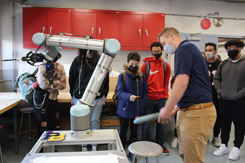 A group of students all wearing masks, watch as one of their teachers, wearing a blue shirt and light tan pants operates a robotic arm.