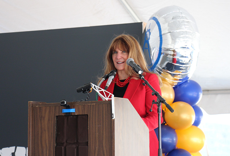 A woman in a red suit and black blouse and shoulder-length blonde hair talks at a podium into a microphone. There are blue and gold balloons behind her.