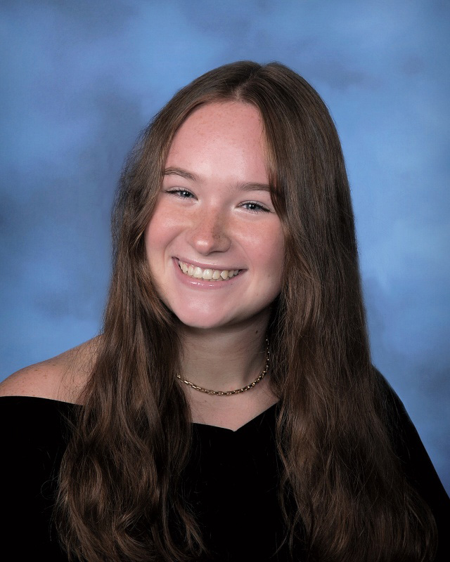 A young woman in a black robe off the shoulders. She has long brown hair, is wearing a gold necklace and is smiling. There is a blue background.