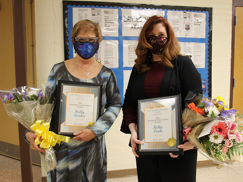 Two women are holding flowers and plaques. The one on the left is wearing a green print dress and a blue face mask. She has short light hair. The woman on the right is wearing a black jacket and maroon shirt. She has long red hair and is wearing a dark mask.