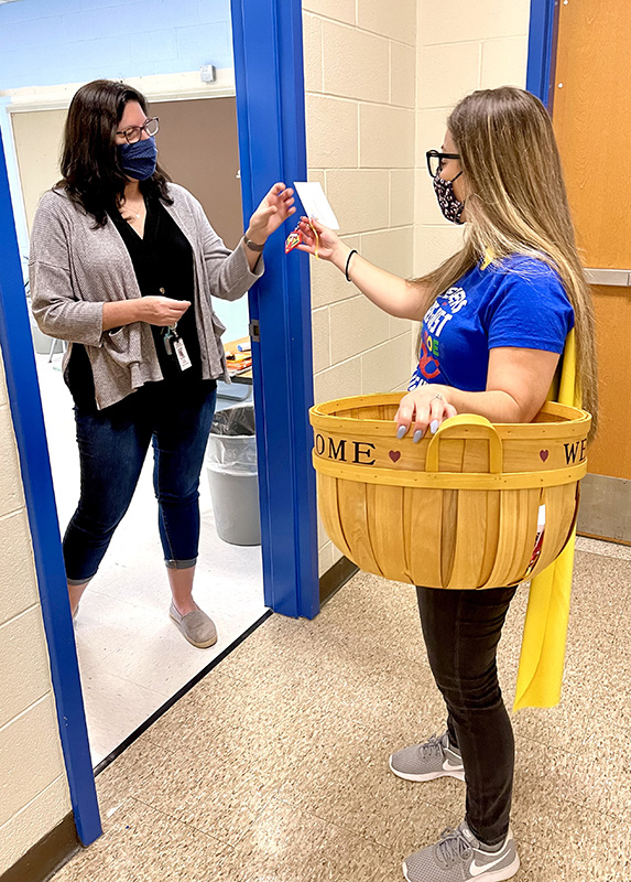 Two women in a doorway. One on the left is wearing black shirt andpants and a cream color sweater. The one on the right is carrying a wicker basket and is wearing a blue shirt and black pants. She has long hair and is handing a key chain to the woman on the left.