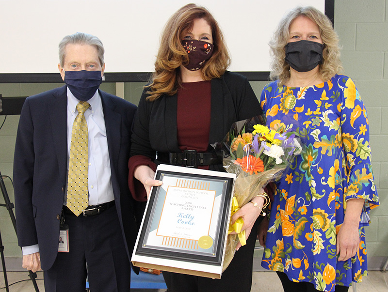Three people are standing for the photo. A man on the left is wearing a suit, gold tie and dark facemask. The woman in the center has long red hair and is weearing a dark facemask and black jacket with maroon shirt. She is holding flowers and a plaque. The woman on the righ tis wearing a blue flowered dress and black mask She has shoulder length blonde curly hair.