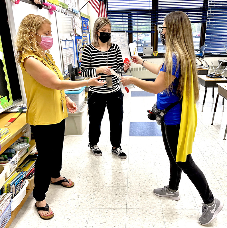 A woman with long hair wearing a gold cape and blue shirt hands a key chain to two women in a classroom. One woman has long blonde hair and is wearing a yellow shirt and black kpants. The woman in the center is wearing a black and white striped shirt and black pants. All are wearing masks.