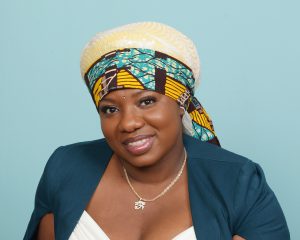 A woman smiling wearing a blue sweater and white shirt and a headcovering of white, blue and yellow.