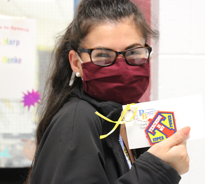 A woman with long dark hair and glasses, wearing a maroon mask, smiles and holds up her key chain that says Teaching is my super power.