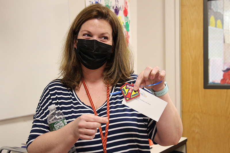 A woman with shoulder-length brown hair wearing a blue and white striped shirt holds up a keychain that says Teaching is my super power along with a small note saying we appreciate all that you do.