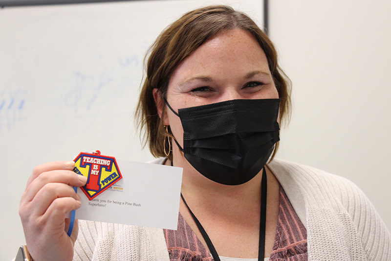 A woman with short brown hair wearing a black facemask, and white sweater holds up her keychain that reads Teaching is my super power.