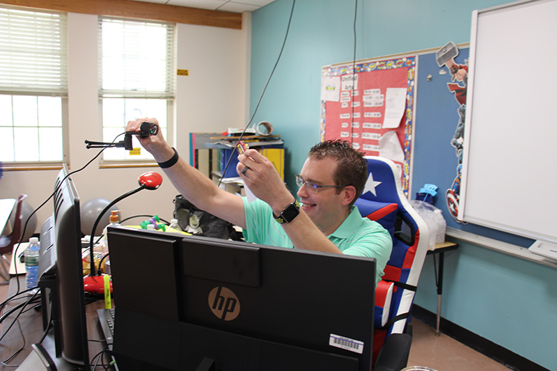A man in a green short-sleeved shirt sits behind a computer and holds up his gift, a keychain that says Teaching is my super power, to his remote students who are watching. He is smiling broadly.