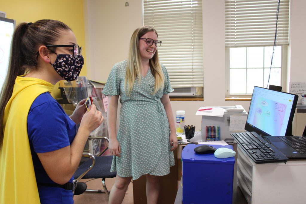 A woman with a blue shirt and yellow cape wearing a mask stands in front of a classroom with a woman in a green dress and long blonde hair. They are talking to a computer screen where a class full of elementary students are watching. 