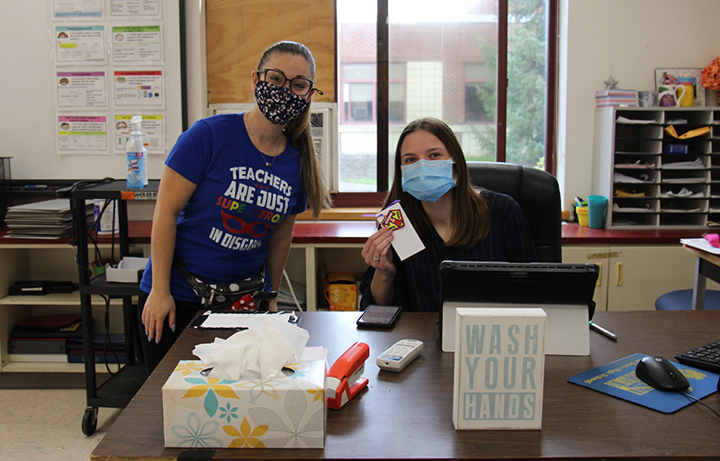 A woman is sitting at her desk, with a compute rscreen in front. She has long dark hair and is wearing a blue mask. She is holding a keychain that was just given to her by the woman standing next to her. She is wearing a blue t-shirt that says Teachers are just super heroes in disguise. On the desk is a sign that says wash your hands.
