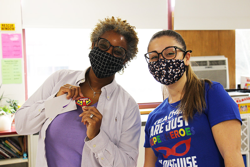 A woman with short hair wearing a blue mask, purple shirt and white button down shirt holds a key chain that says Teaching is my super power. Next to her is a woman with glasses, ponytail, mask and a blue shirt that says Teachers are just super heroes in disguise.