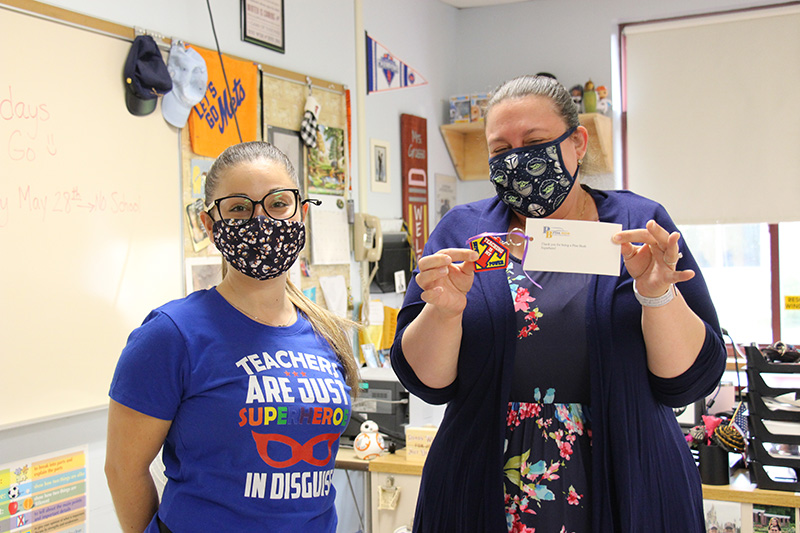 Two women in a classroom. The one on the left is wearing a blue t shirt that says Teachers are just super heroes in disguise. The woman on the right has her hair pulled back, wearing a blue flowered dress and sweater, holding up the keychain she just received. It says Teaching is my super power. Both are wearing face masks.