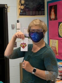 A woman with short blonde hair holds up her key chain that says Teaching is my super power. She is wearing a face mask and a green shirt.