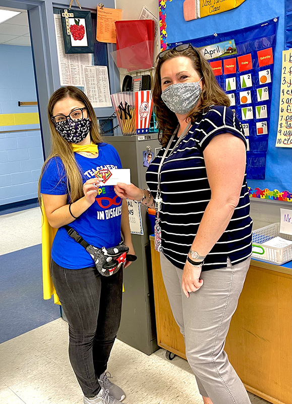 A woman with long hair, wearing a blue shirt and gold cape hands a key chain gift to a woman wearing light pants and a blue and white striped shirt. Both are wearing masks.