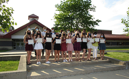 Nineteen high school students stand on steps in front of a high school building with red roof and trees behind them.