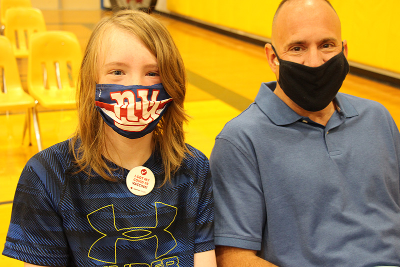 A young man with long blonde hair wearing a blue shirt and mask with the NY Giants logo wears a pine that says I got my COVID-19 vaccine. Next to him is a man with a blue shirt, black mask and very short hair. They are both sitting in a gymnasium.