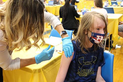 A young man with long blonde hair, wearing a blue tshirt and a blue mask with a red and white NY Giants logo, gets a vaccine by a woman with mid-length light hair wearing blue gloves.