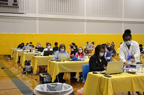 Several tables with yellow tablecloths lined up. There are adults at each of them and a student at each also. The adults have laptops in front of them. It is in a large gymnasium.