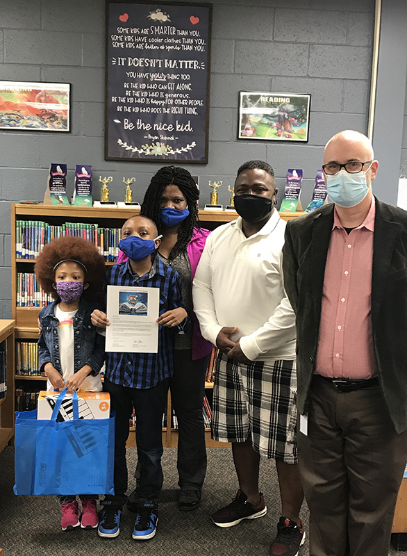 A little girl stands holding a bag and a young boy holds a certificate. Their parents are gehind them and another man is standing on the right.