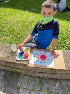 An elementary school boy wearing a blue and blackshirt sits on a stone bench painting. He is wearing a green mask. It's a bright sunny day.